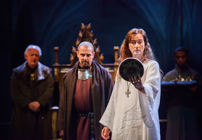 Long-haired Richard in wrinkled white robe and gold cross on a chain around his neck looks into a round mirror he holds with his left hand as, behind him, Bolingbrok in armored breast plate and orange tunic stares off in irritation, and furth in the bacgkround we see York, a page holding a pillow with crown and scepter, and the throne.