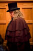 Scrooge next to a wood wall with audience around him, he's wearing a brown multi-layered topcoat and a black tophat