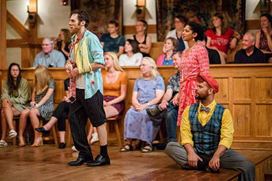 Production photo by Marek K Photography of a bedraggled and wounded Antipholus of Ephesus along with his Dromio (sitting on the edge of the stage) and his wife Adriana addressing the Duke (off-camera) with the audience in the background.