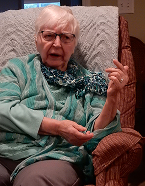 Carol Kelly wearing a green sweater and glasses and expressing herself with hand gestures, sits in an overstuffed brown chair with a tany blanket hung over its back.