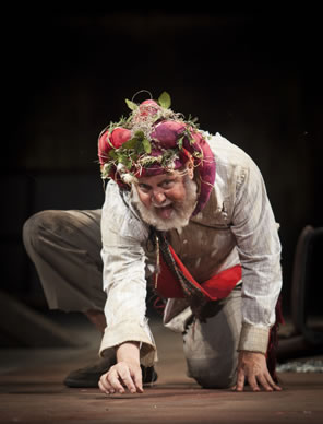 Lear, one knee on the ground, picking at the flor with his right fingers, wearing dirty white shirt and pants, his red regal sash, and the Fool's coxcomb covered in brambles