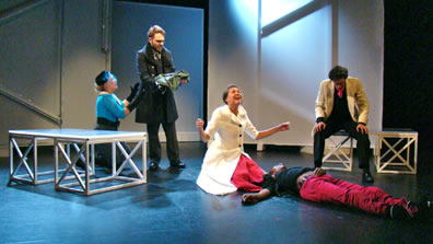 On a stage of scrim panels and metal tables, Lady Montague kneels beside the Prince looking down at Mercutio's kilt, Lady Capulet in long white coat and pink dress kneels over the prone body of Tybalt as Capulet sits on one of the tables next to the body.