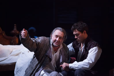 Henry in bedclothes and robe on the floor next to his used bed points in the distance as Hal kneeling beside him in vest and white shirt holds his arm.