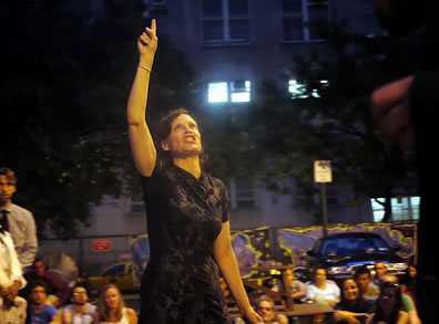 Margaret in shape-forming black print dress, right arm pointing up as she curses to heaven